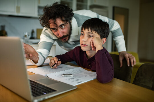 Father helping bored son with homework on laptop at kitchen table
