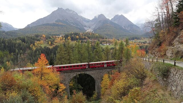 A classic red Swiss train glides across a rustic stone viaduct nestled in golden autumn forests, with the historic Tarasp Castle perched beneath towering, snow-dusted alpine peaks.