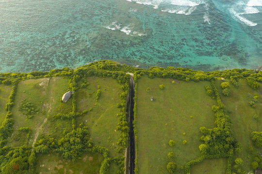 Aerial view of the road at Pantai Tanah Barak carved through limestone cliffs leading to the turquoise ocean, surrounded by lush green fields South Kuta, Bali, Indonesia.