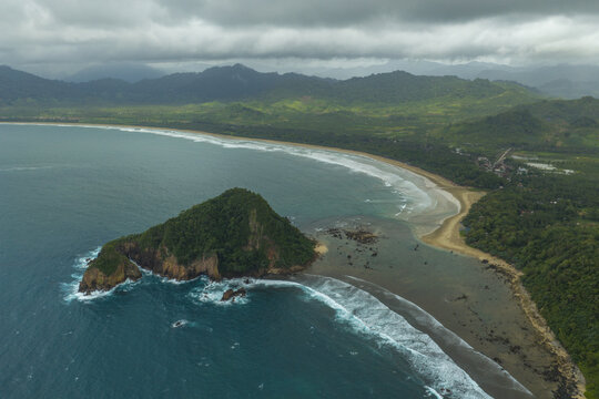 Aerial view of Red Island Beach with its rocky island, sandy coastline, and lush green hills under a cloudy sky in East Java, Indonesia.