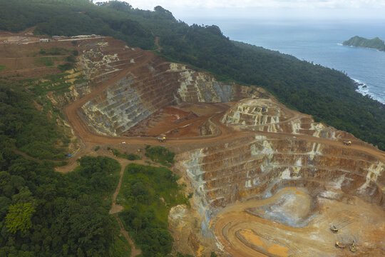 Aerial view of the Tujuh Bukit gold and copper mine with terraced excavations, mining trucks, and surrounding forest near the coastline in Kecamatan Pesanggaran, Jawa Timur, Indonesia.