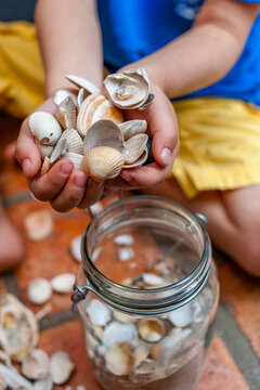 A boy holds a seashell collection over a glass jar outdoors in Sylt