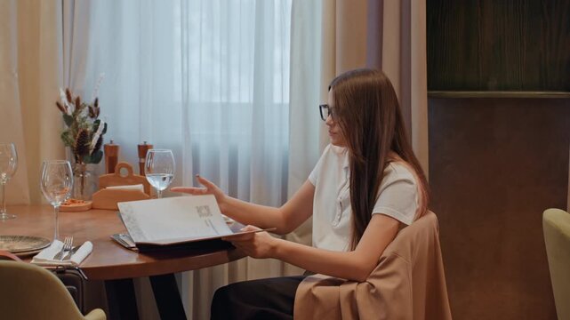 Caucasian woman seated at dining table reviewing menu and adjusting glasses, solo diner flips pages and studies choices as freelancer and food critic warm interior with vase, curtain, wineglass, plate