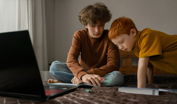 Teenager and boy study together with a laptop and textbook at home