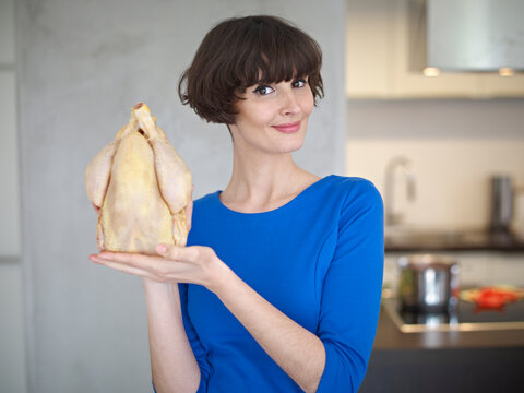 Germany, Cologne, Young woman with raw chicken in kitchen, smiling, portrait