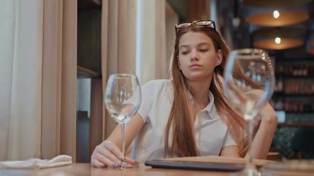 Young woman seated at restaurant table, quiet evening in cozy bistro with sunglasses on head and long braids, empty wine glass in foreground, subtle window reflections, soft bokeh lights, cinematic