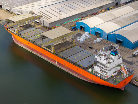 Aerial view of a large orange cargo ship docked at an industrial port facility with warehouses and storage areas in United Kingdom.