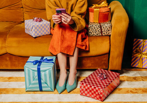 Stylish woman sits on a couch with a gift and uses a smartphone