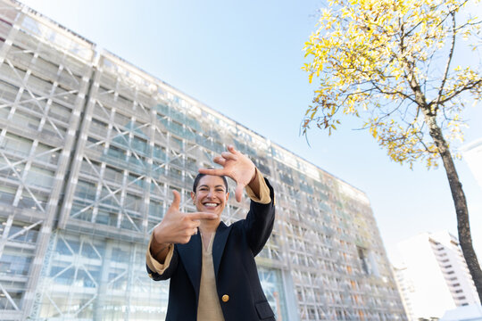 Smiling businesswoman frames view with hands outdoors at office plaza