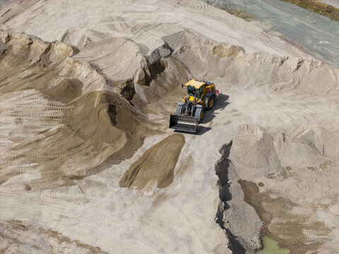 Aerial view of a yellow wheel loader working in a large sand and gravel quarry with piles of earth and tire tracks on the ground in the United Kingdom.
