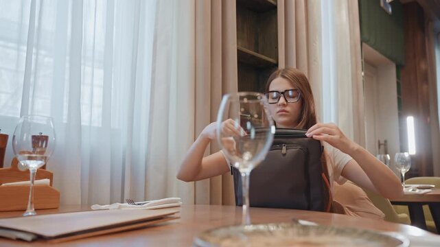 Young woman securing laptop bag before leaving restaurant, zipping closure and straightening glasses, calm confidence and warm interior, glassware and place setting on table, end of work from cafe