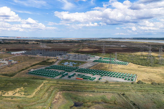 Aerial view of a large battery energy storage system facility with rows of green containers, power infrastructure, and transmission towers in a rural landscape in the United Kingdom.