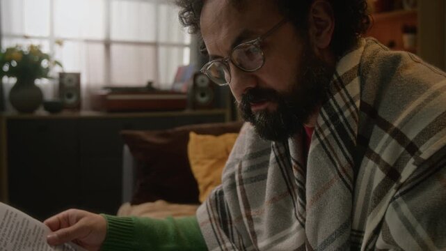 Middle aged Caucasian man with glasses reading industrial technology newspaper at home, wrapped in blanket. Low angle tilt up shot is ideal for print media campaigns or financial literacy ads