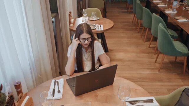 White woman working alone in cafe, on laptop rubbing temples, deadline tension, warm wood interior with green chairs and table settings, coffee cup and notepad nearby, subdued lighting creating