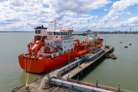 Aerial view of a large orange tanker ship docked at an industrial pier with pipelines and storage facilities in the background under a bright blue sky in the United Kingdom.