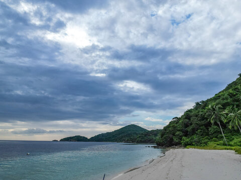 Aerial view of Linapacan Island featuring a pristine white sand beach, lush tropical hills, and clear turquoise waters under a dramatic cloudy sky in Linapacan, Mimaropa, Philippines.