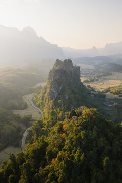 Aerial view of Nam Xay Viewpoint surrounded by lush green forests, winding roads, and agricultural rice fields in the golden light of the morning in Naka, Vientiane Province, Laos.