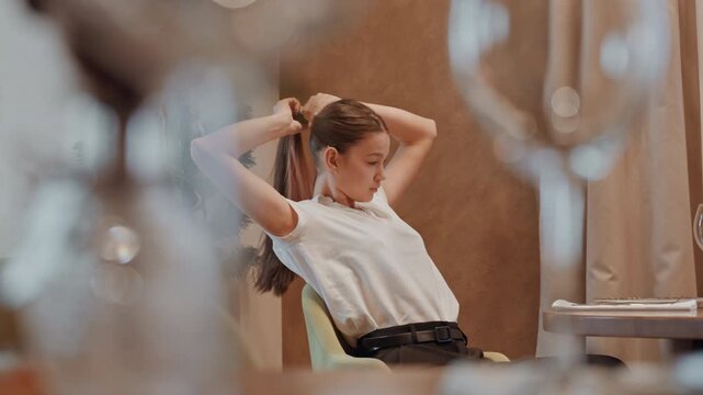Young white woman in restaurant chair, styling hair through reflection, preparing for evening engagement, blurred glassware foreground, candid motion, tailored blouse, confident expression, editorial