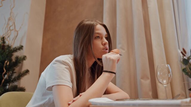 Young black woman tasting pastry, braided hair and playful smile, soft curtain light, small pine plant and glass on table, expectant gaze while holding treat, candid reaction and savoring moment, cozy