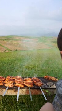 Young boy watching chicken wings shashlik grilling on skewers over a mangal with a scenic green valley background, slow motion vertical video.