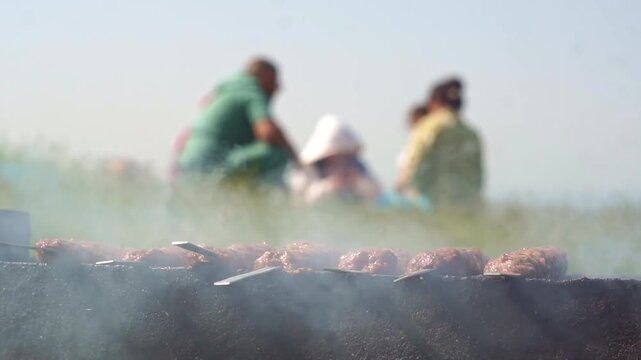 Traditional lula kebab shashlik grilling on a mangal with smoke, blurred family enjoying a picnic in the background, horizontal video.