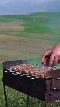Close up of a man turning skewers of traditional minced meat lula kebab shashlik on a mangal grill with a scenic green mountain background, vertical video.