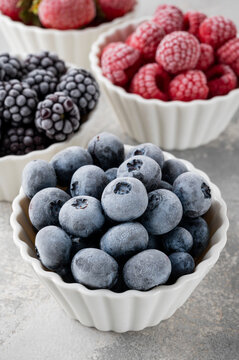 Assorted frozen berries in white bowls: strawberries, blueberries, raspberries, and blackberries on a light stone background. Healthy food. Close up