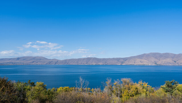 Calm blue waters of Lake Sevan with autumn trees in the foreground and a mountain range under a clear blue sky, Armenia.