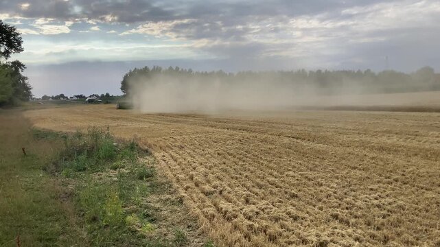 Combine Harvester Turning at Field Edge, Continuing Wheat Harvest While Dust Rises and Tractor with Grain Trailer Tractor Departs in Golden Evening Light