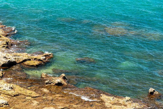 Turquoise sea water lapping over rocky coastal shoreline on sunny day, clear ocean waves with visible underwater rock formations and vibrant teal color