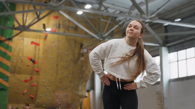 White woman stretching in climbing gym, hands on hips, braid, colorful climbing holds, high ceiling and industrial lighting, focused breath and mobility work, casual sweatshirt and leggings
