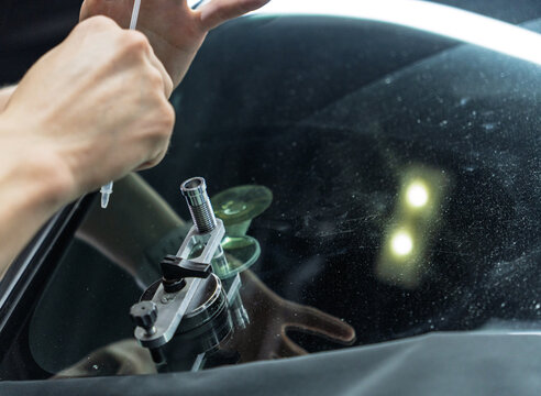 Professional technician uses a special tool and liquid polymer for removing chips and cracks on a car windshield. Close up of hands during a careful vehicle window restoration process. 