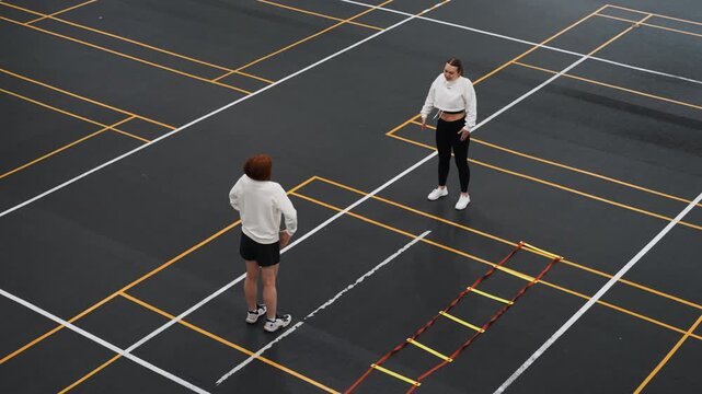 Coach demonstrates shuttle placement and tactics on marked court, instructional dialogue and slow demonstration of angles, attentive trainee watches amid agility ladder and court lines