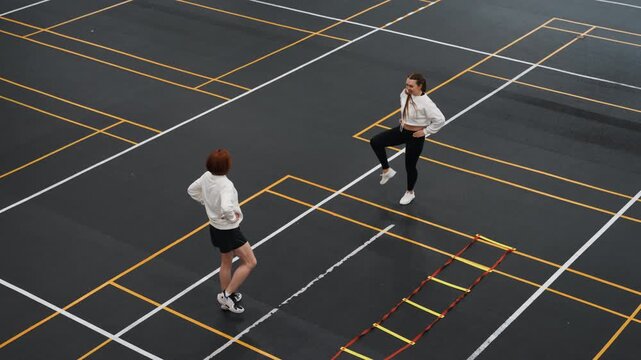 Two players in casual badminton rally rooftop, playful exchange of serves and footwork across marked pavement, friends laughing while practicing light drills near agility ladder with urban skyline