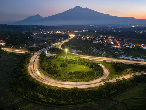 Aerial view of Jalur Exit Tol Salatiga at twilight, featuring the curved highway ramp surrounded by rice fields, mountain silhouettes, and city lights in Tengaran, Central Java, Indonesia.