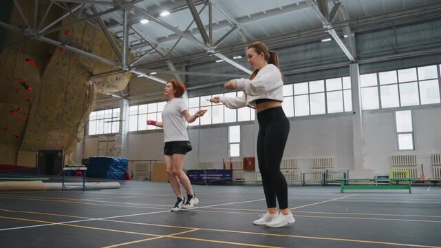 Two athletes warming up on indoor court practicing badminton serves and technique drills with coachlike instruction, deliberate footwork and timing exercises, climbing wall and high ceiling backdrop,