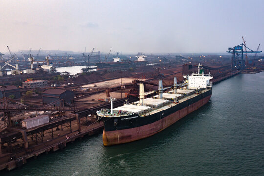 Aerial view of a large cargo ship docked at Paradip Port, surrounded by industrial cranes, loading infrastructure, and piles of raw materials in Paradeep, Odisha, India.