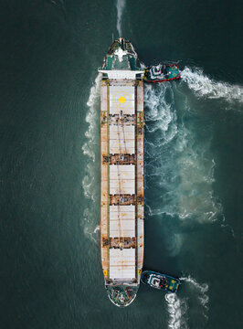 Aerial view of a large cargo ship being maneuvered by two tugboats in the waters of Paradip Port, showing the vessel's deck and the surrounding sea wake in Paradeep, Odisha, India.