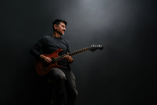 M&uacute;sico guitarrista en penumbra, sonriendo y sentado con actitud feliz, ganadora y heroica con su instrumento musical naranja. Fotograf&iacute;a emotiva de alto contraste.