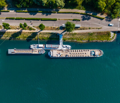 Aerial view of river cruise ships docked at a pier along the Rhine river with a nearby road and lush green trees in Vogelgrun, Grand Est, France.