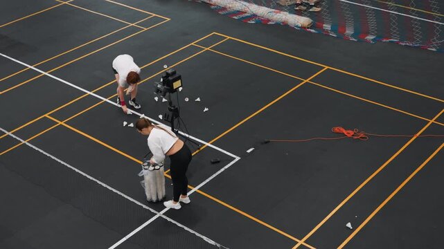 Badminton court training session with camera. Two staff adjust shuttle basket and tripod while preparing practice drill on marked indoor court, scattered shuttlecocks, focused atmosphere, slow motion