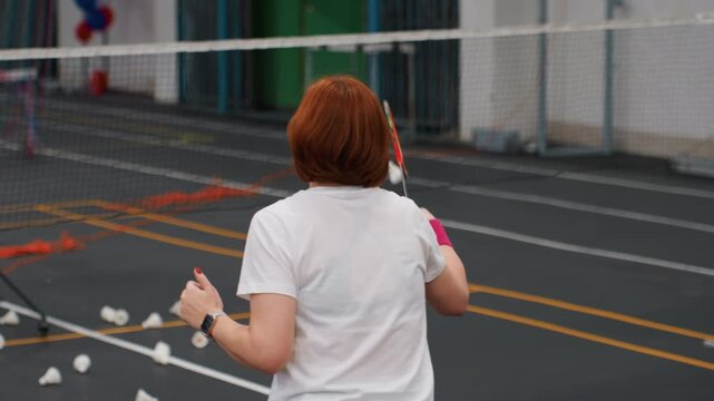 White women practicing badminton rally together on indoor court with scattered shuttlecocks, red hair player and ponytailed partner exchanging fast volleys, footwork drills, dynamic movement and coach
