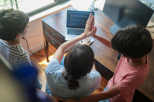 Family at home using a laptop, a mother and two boys high-fiving over a desk, celebrating success and teamwork while learning to code on a computer