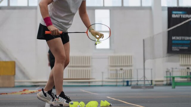 White woman serving badminton in indoor sports hall, coach trainee practicing accuracy, rackets and pile of shuttlecocks, measured tosses and concentrated stance, bright windows and methodical