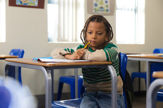 African American boy sitting at wooden desk holding pencil near mouth, wearing striped sweater
