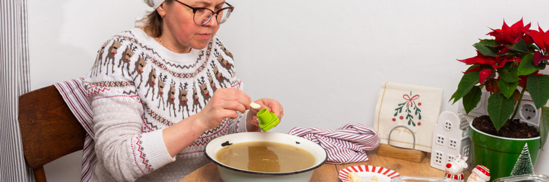In a kitchen in Ukraine, a woman is making kholodets, aspic, or jellied meat. She grates garlic into a bowl of broth. On the table, there are other ingredients and containers for the dish
