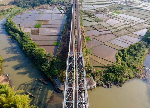 Aerial view of a double railway bridge crossing a river, flanked by lush green and flooded rice paddies in Banyumas Regency, Central Java, Indonesia.