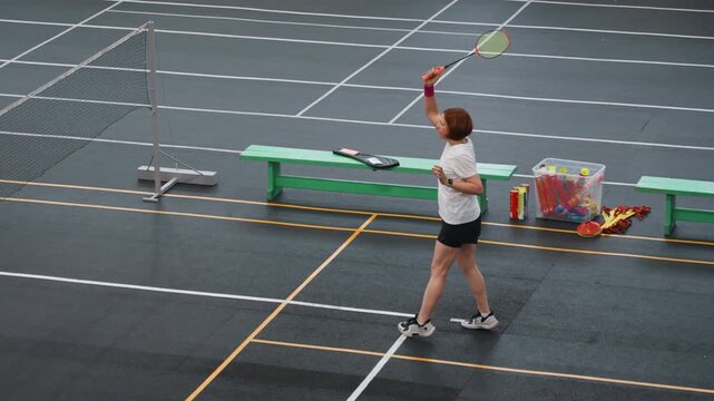 Recreational badminton serve practice on court with solo player rehearsing serves and returns, casual club setting, benches with equipment, measured swings and shuttlecock arc under gym lights