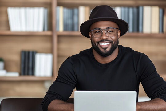 Friendly black male blogger with hat smiles at camera, while sitting at laptop in home office with book-lined shelves