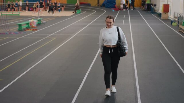 Young caucasian woman walking indoor court confident stride with black bag over shoulder, white hoodie, black leggings and sneakers wide sports hall with lane markings, equipment and spectators
